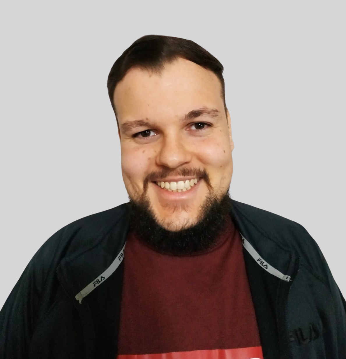 Foto de Rodrigo Molter sorrindo enquanto olha para a câmera. Rodrigo é um homem, com rosto oval e possui barba. Utiliza uma camisa bordo e um casaco preto. A foto possui um fundo cinza.
